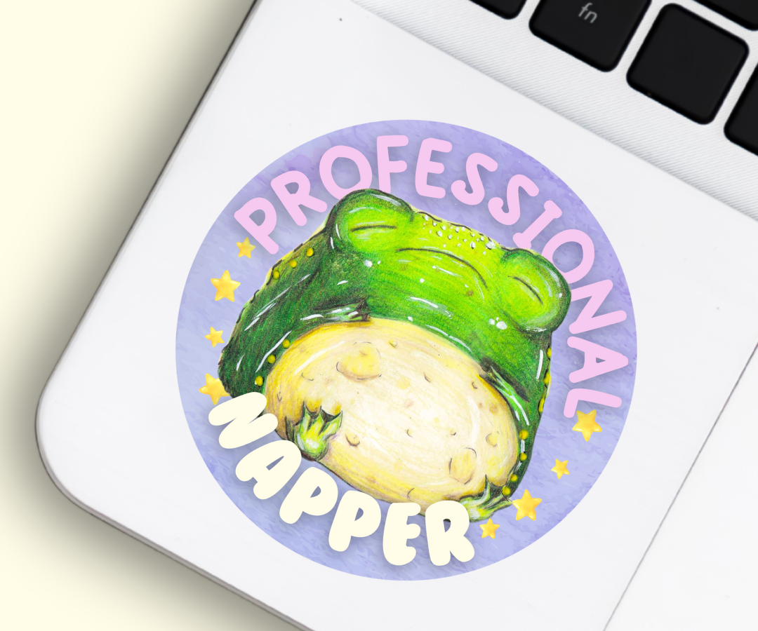 image shows a round purple vinyl sticker, featuring a chubby sleeping toad, around him are the words 'professional napper' and he is surrounded by stars either side of him. The sticker is placed in the left bottom corner of a silver laptop, beside the mousepad