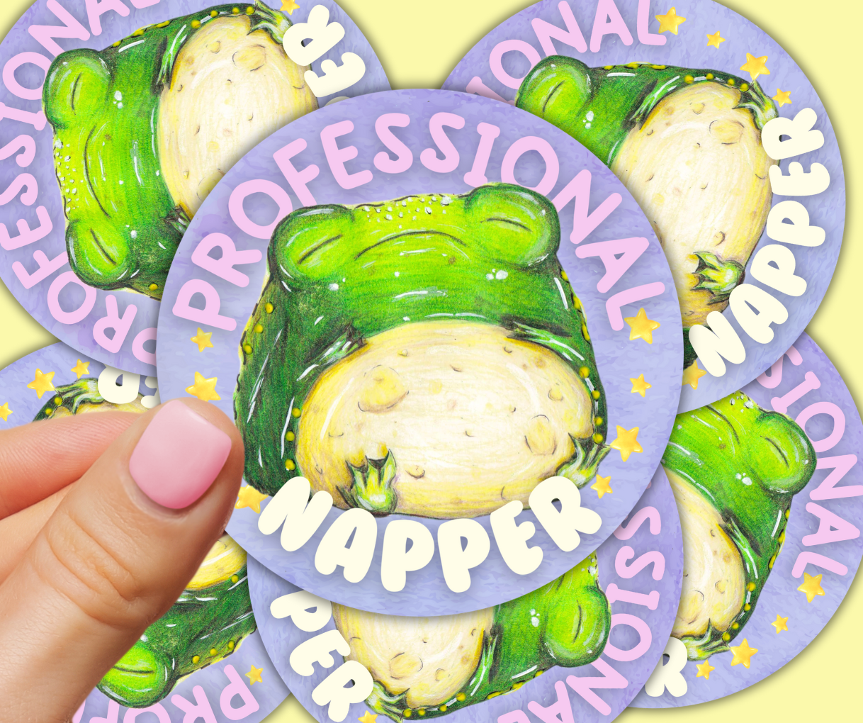 image shows a round purple vinyl sticker, featuring a chubby sleeping toad, around him are the words 'professional napper' and he is surrounded by stars either side of him, the sticker is being held between two fingers, in front of a scattered pile of the same sticker underneath