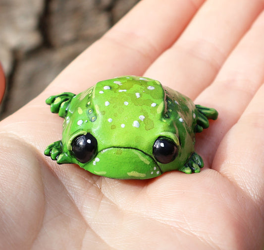 image shows miniature glumbert, a sad toad who is placed in the palm of someones hand. he is round and flat in shape, with big black cute eyes, and a downturned mouth, he has wide feet which splay from underneath his body. he is green with an assortment of white, brown and yellow spots