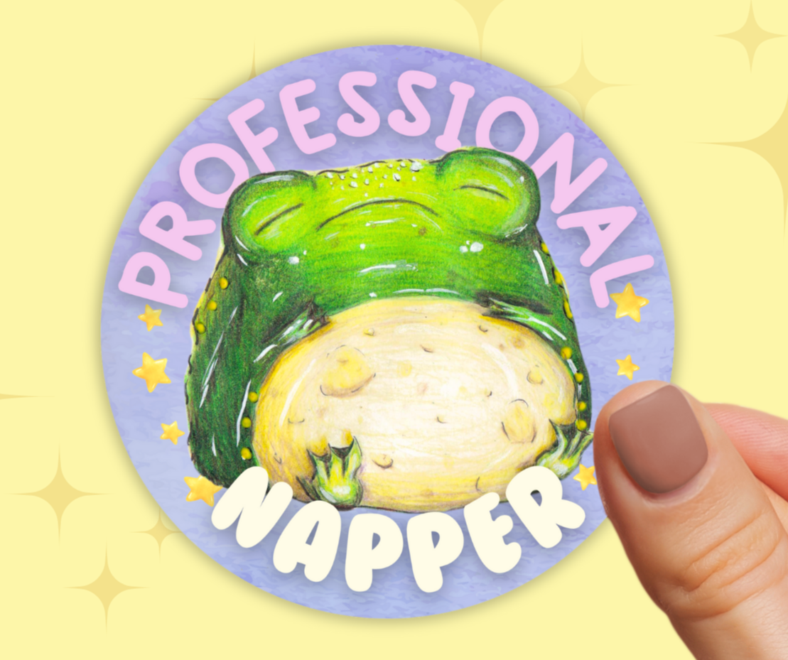 image shows a round purple vinyl sticker, featuring a chubby sleeping toad, around him are the words 'professional napper' and he is surrounded by stars either side of him. the sticker is being held in between two fingers in front of a pastel yellow background