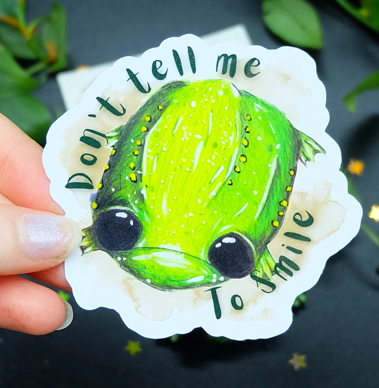 Image shows Glumbert the sad toad, a green spotted toad with a sad mouth and big black eyes. He is placed upon a brown watercolour marking with the words 'Don't tell me to smile' in dark green. The sticker is held between a finger and thumb over a dark background
