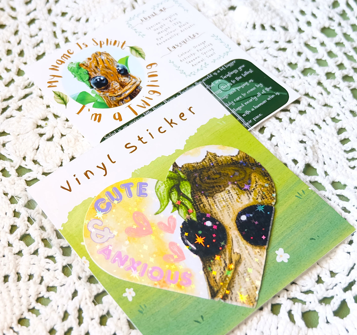 Image shows the packaged vinyl sticker included in the Splint product, as well as the character backstory card. They are laid out at an angle upon a cream doilie which is placed on a green table