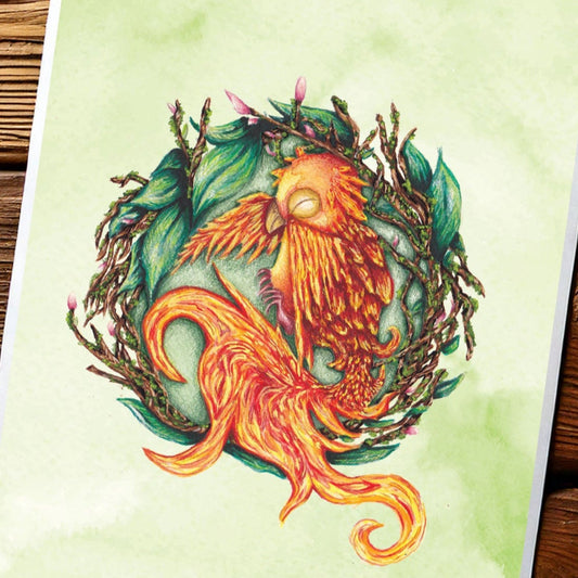 image shows a portrait art print featuring a painting of a baby phoenix resting on it's side, inside a nest made of twigs, leaves and budding flowers. behind the main detail of the painting, there is a green watercolour wash making the background mottled green. the paper is placed on the top of a dark wooden antique table