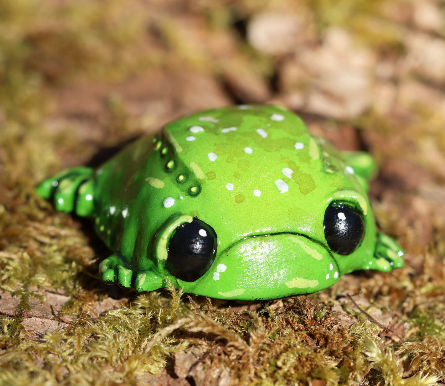 image shows miniature glumbert, a sad toad . he is round and flat in shape, with big black cute eyes, and a downturned mouth, he has wide feet which splay from underneath his body. he is green with an assortment of white, brown and yellow spots