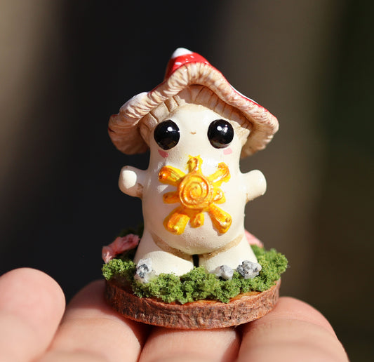 the photo shows alba, the light bringer, who is a fly agaric mushroom. she has a golden sunshine on her soft belly, big cute black eyes, a red and white spotted cap, and she stands with her arms outspread in a loving stance. she is attached to a circular wooden base which is decorated with decorative moss, and small stones. the sculpture is placed on the finger tips of an outstretched hand