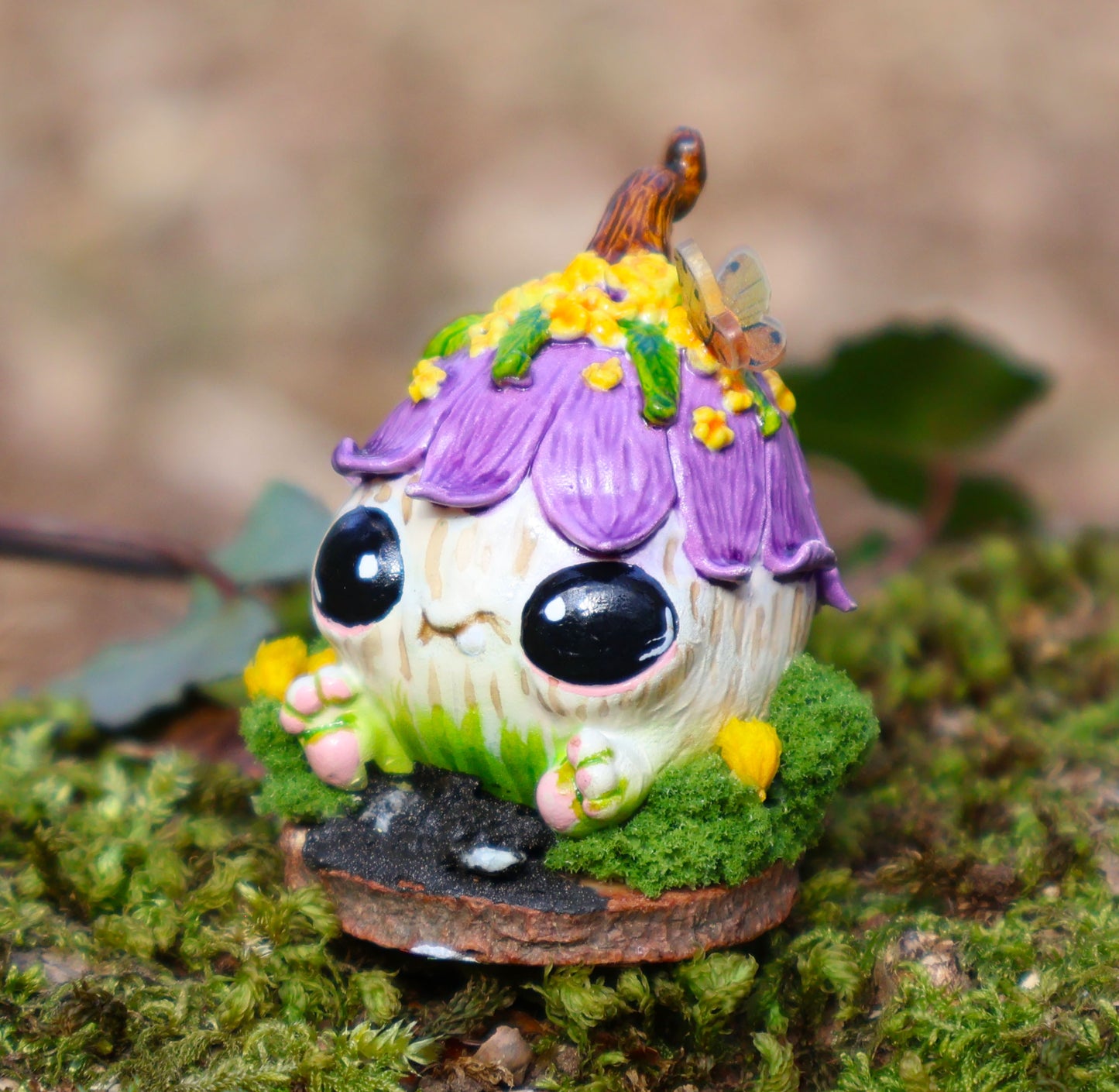 Image shows Blush, the mischievous sprouting, She is seated on a wooden circular base, surrounded by moss & yellow flowers in front of her, details look like mud and rocks. Blush is a round with little feet sticking out from the bottom of her body, she has no arms, two huge black adorable eyes, & a cheeky smile. her body is white & green she has a purple flower for a hat, with yellow flowers, green leaves and a brown stem, she has a butterfly perched in one of her flowers