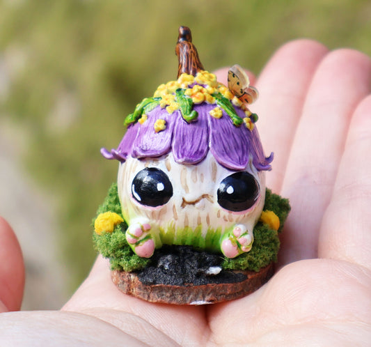 Image shows Blush, the mischievous sprouting, held in the palm of someone's hand. She is seated on a wooden circular base, surrounded by moss & yellow flowers in front of her, details look like mud and rocks. Blush is a round with little feet sticking out from the bottom of her body, she has no arms, two huge black adorable eyes, & a cheeky smile. her body is white & green she has a purple flower for a hat, with yellow flowers, green leaves and a brown stem, she has a butterfly perched in one of her flowers