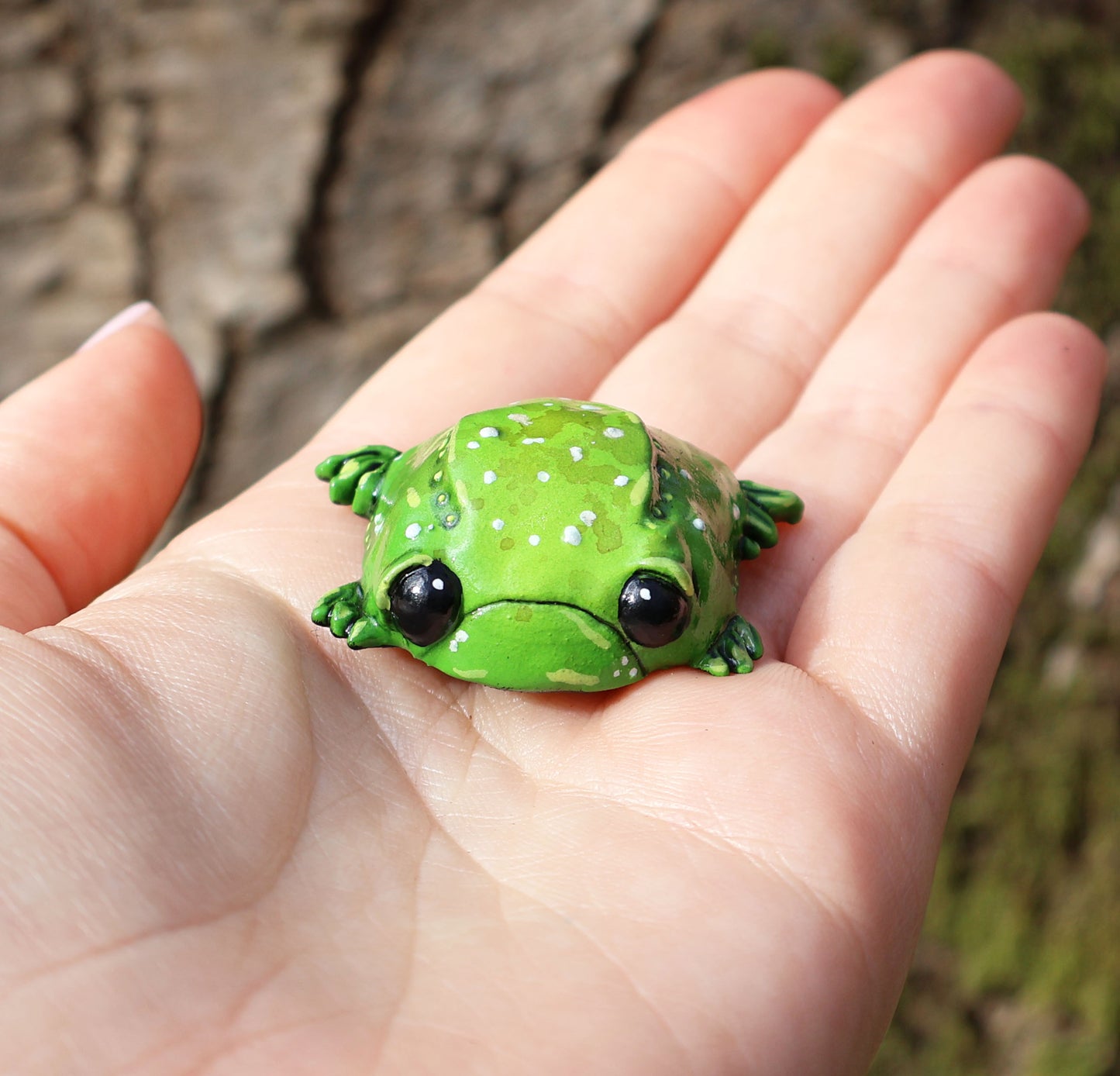 image shows miniature glumbert, a sad toad who is placed in the palm of someones hand. he is round and flat in shape, with big black cute eyes, and a downturned mouth, he has wide feet which splay from underneath his body. he is green with an assortment of white, brown and yellow spots