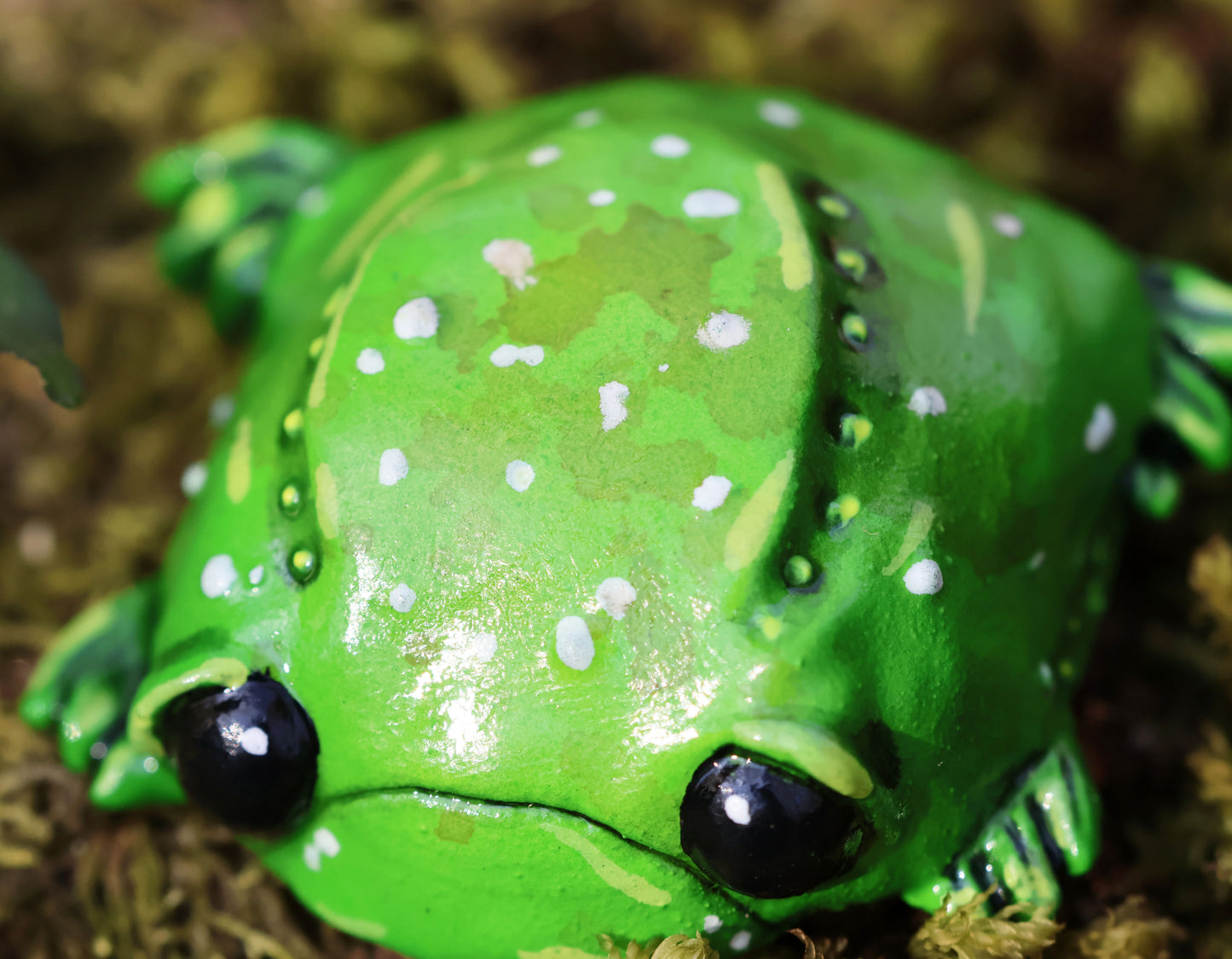 image shows miniature glumbert, a sad toad he is round and flat in shape, with big black cute eyes, and a downturned mouth, he has wide feet which splay from underneath his body. he is green with an assortment of white, brown and yellow spots