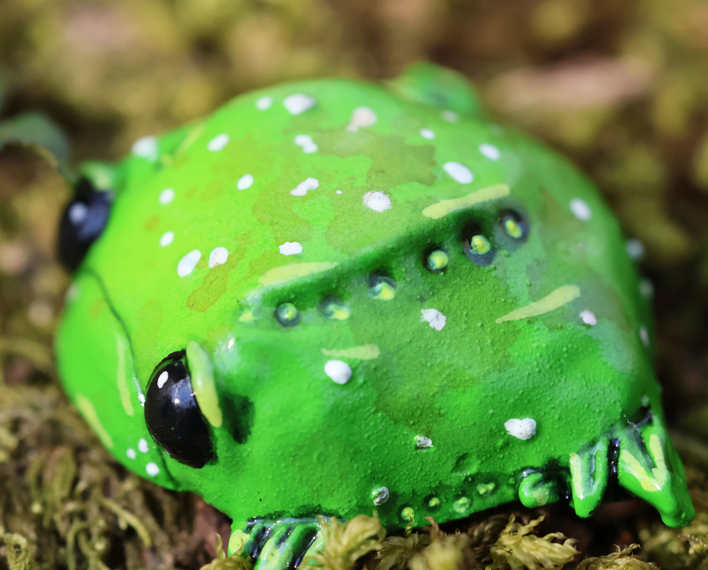 image shows miniature glumbert, a sad toad. he is round and flat in shape, with big black cute eyes, and a downturned mouth, he has wide feet which splay from underneath his body. he is green with an assortment of white, brown and yellow spots