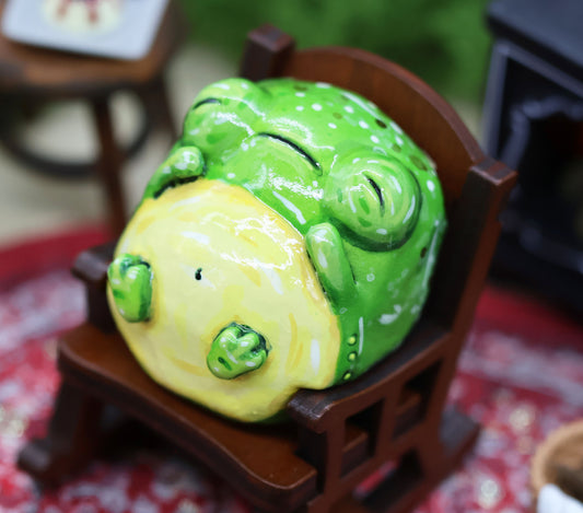 image shows a handpainted figurine of a sleeping toad, Glumbert who is leaning back in a miniature wooden rocking chair. Glumbert has his arms resting on his round stomach, and his feet are up in a relaxed position. His eyes are closed, and his mouth as slightly downturned as he sleeps. the rocking chair is placed upon a red patterned rug, and in the blurred background you can see a black wood burning stove, and a wooden side table