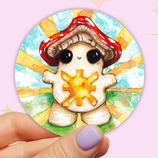 Image shows a round vinyl sticker, being held between a finger and thumb in front of a baby pink background. The sticker features a happy smiling chubby toadstool mushroom, arms wide open with a warm sunshine on her belly. Behind her are rays of golden light covering a watercolour impression of green grass and blue sky.