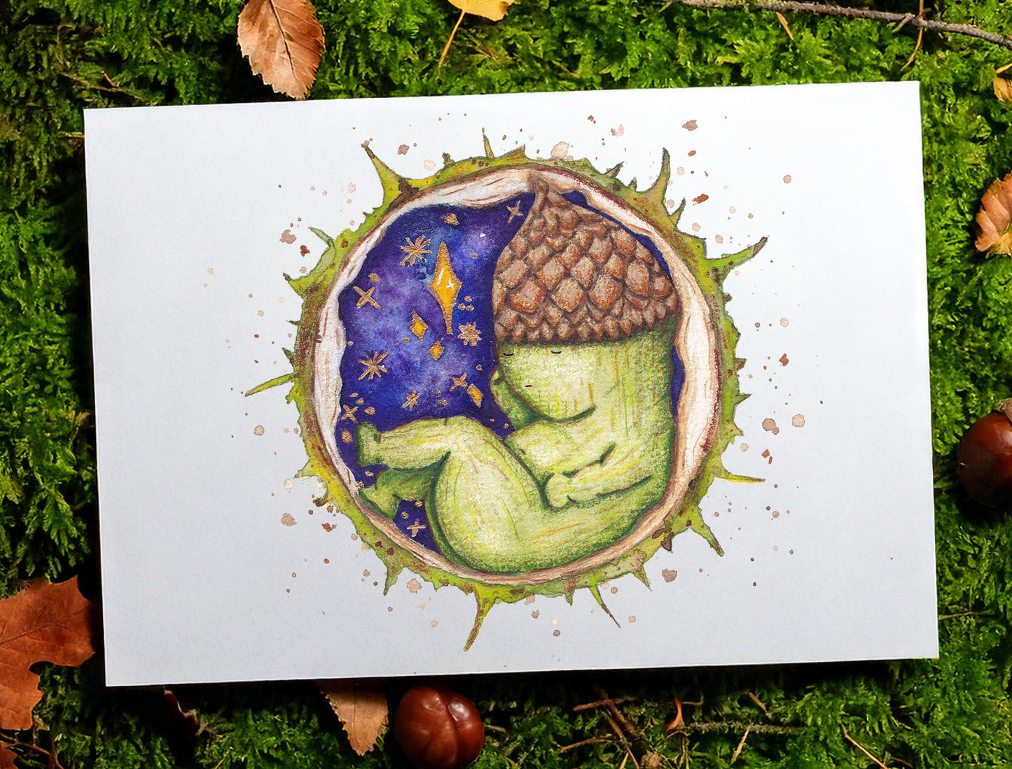 image shows an art print propped against a mossy tree. the art print features a small acorn baby creature sleeping inside the shell of a conker. behind them is a magical starry night