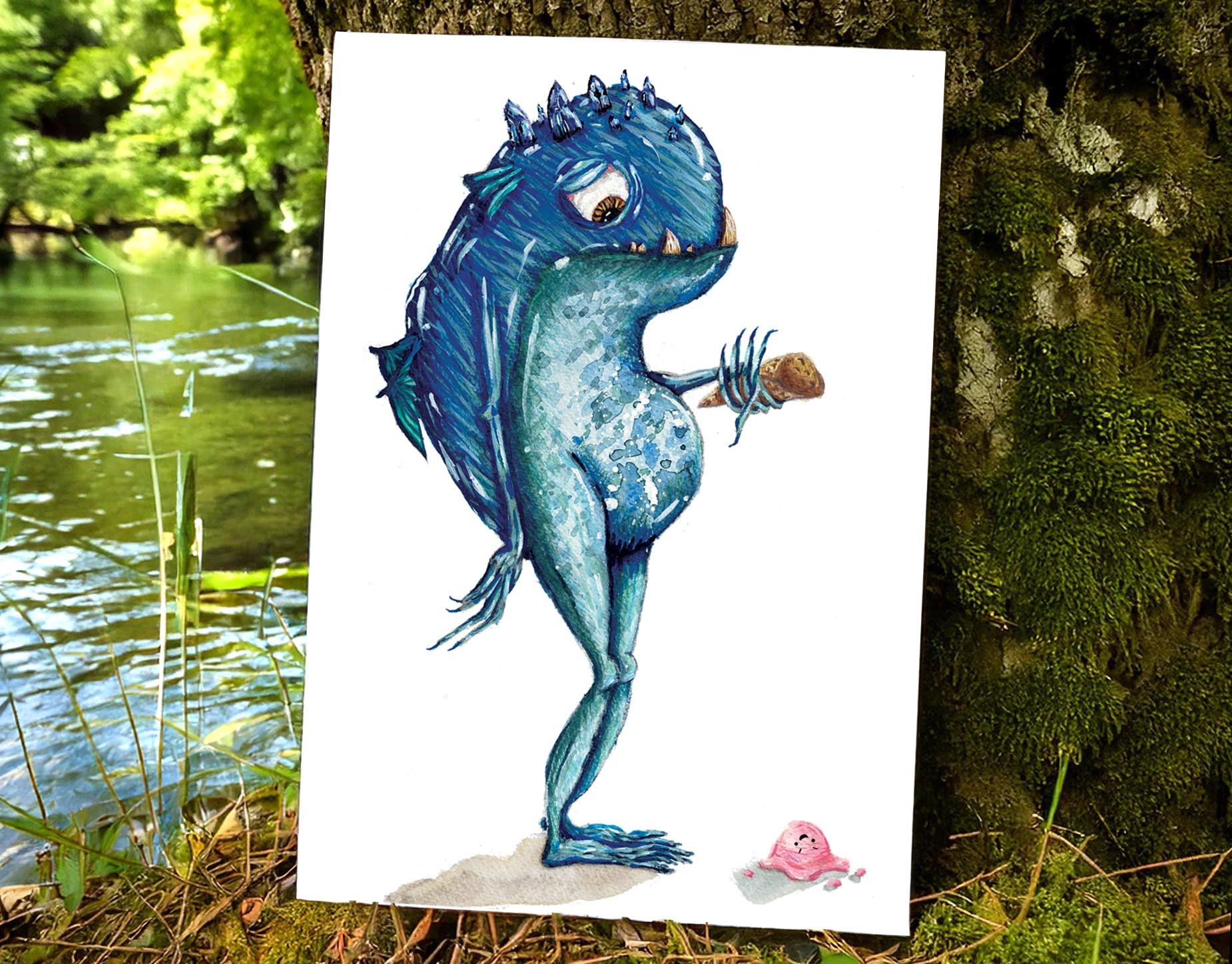 art print placed on a mossy riverbank. art print features a mystical fantasy creature, who is blue, with small wings and crystal points coming from their head. he looks down hopelessly at the ice cream that has just fallen to the ground from his cone.