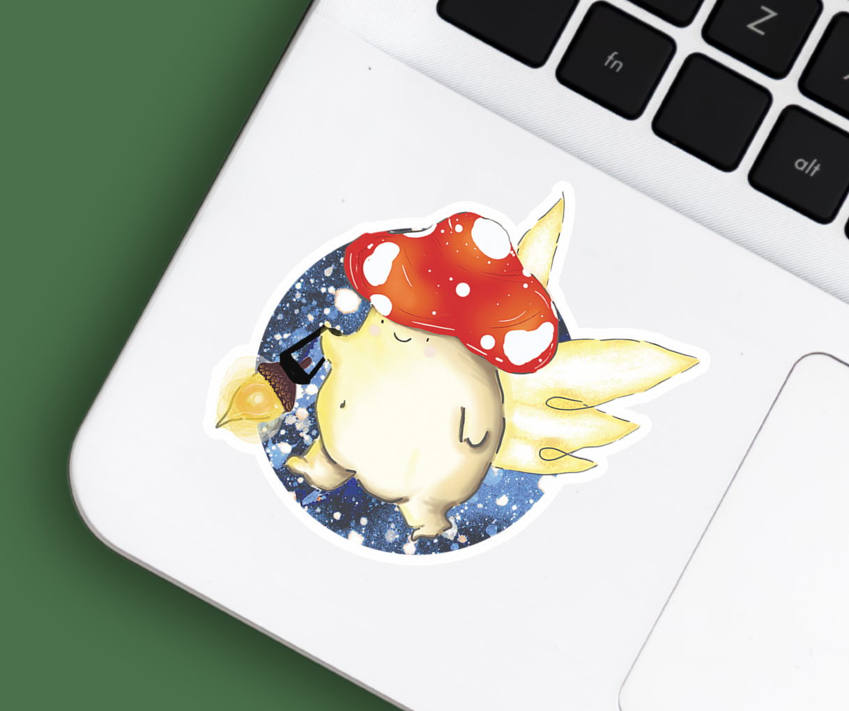 a vinyl sticker featuring a fairy toadstool mushroom with wings, flying through the night sky with a lantern, positioned beside the mousepad of a silver laptop