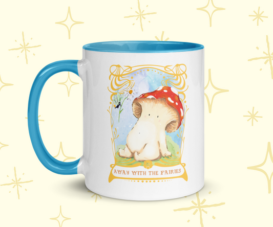 image shows a white ceramic mug which is blue on the inside, and the handle. The design features an illustration of a toadstool mushroom sitting down on mossy grass. she is being handed a daisy by a fairy, the sky is blue behind them. The illustration is framed with an ornate gold art nouveau style framing, with the words at the bottom reading 'away with the fairies'. The mug is imposed in front of a cream background with star graphic details