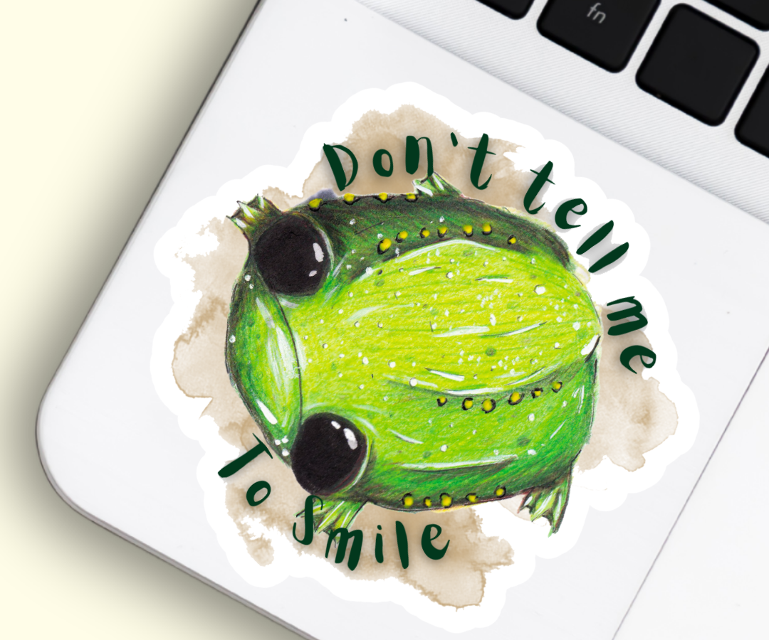 Image shows a sticker placed beside the mousepad of a silver laptop. The sticker features an illustration of Glumbert the Sad  Toad, Glumbert is green with white and yellow spots, he has big black eyes and a sad downturned mouth. Around his body is a brown watercolour smugde and the words, "Don't tell me to smile"