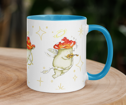 image shows a white ceramic mug which is blue on the uinside, and the handle. The design features illustrations of cherub mushrooms playing instruments and flying amongst a starry backdrop. The mug is placed on top of a wooden log slice with blurred impressions of trees in the background