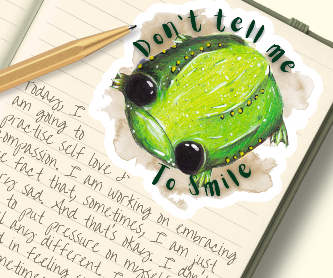 image show a vinyl sticker placed within a journal. The sticker features an illustration of Glumbert the Sad  Toad, Glumbert is green with white and yellow spots, he has big black eyes and a sad downturned mouth. Around his body is a brown watercolour smugde and the words, "Don't tell me to smile"
The journal entry is themed around depression and embracing your sad feelings.