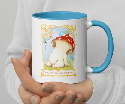 image shows a white ceramic mug which is blue on the inside, and the handle. The design features an illustration of a toadstool mushroom sitting down on mossy grass. she is being handed a daisy by a fairy, the sky is blue behind them. The illustration is framed with an ornate gold art nouveau style framing, with the words at the bottom reading 'away with the fairies'. The mug is being held in two cupped hands by a person wearing a white top
