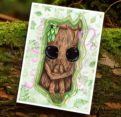 Art print photographed against a mossy tree trunk within a forest. The art print features cute & anxious Splint the Twigling, a wooden fantasy creature who is hugging their knees in a seated position. They have big black eyes & are painted onto a green background with green leaves surrounding them. Pink words read, 'Cute and Anxious' to the left of Splint, & to the right of them are a selection of pink hearts in various sizes. Splint is surrounded by a green & golden glow
