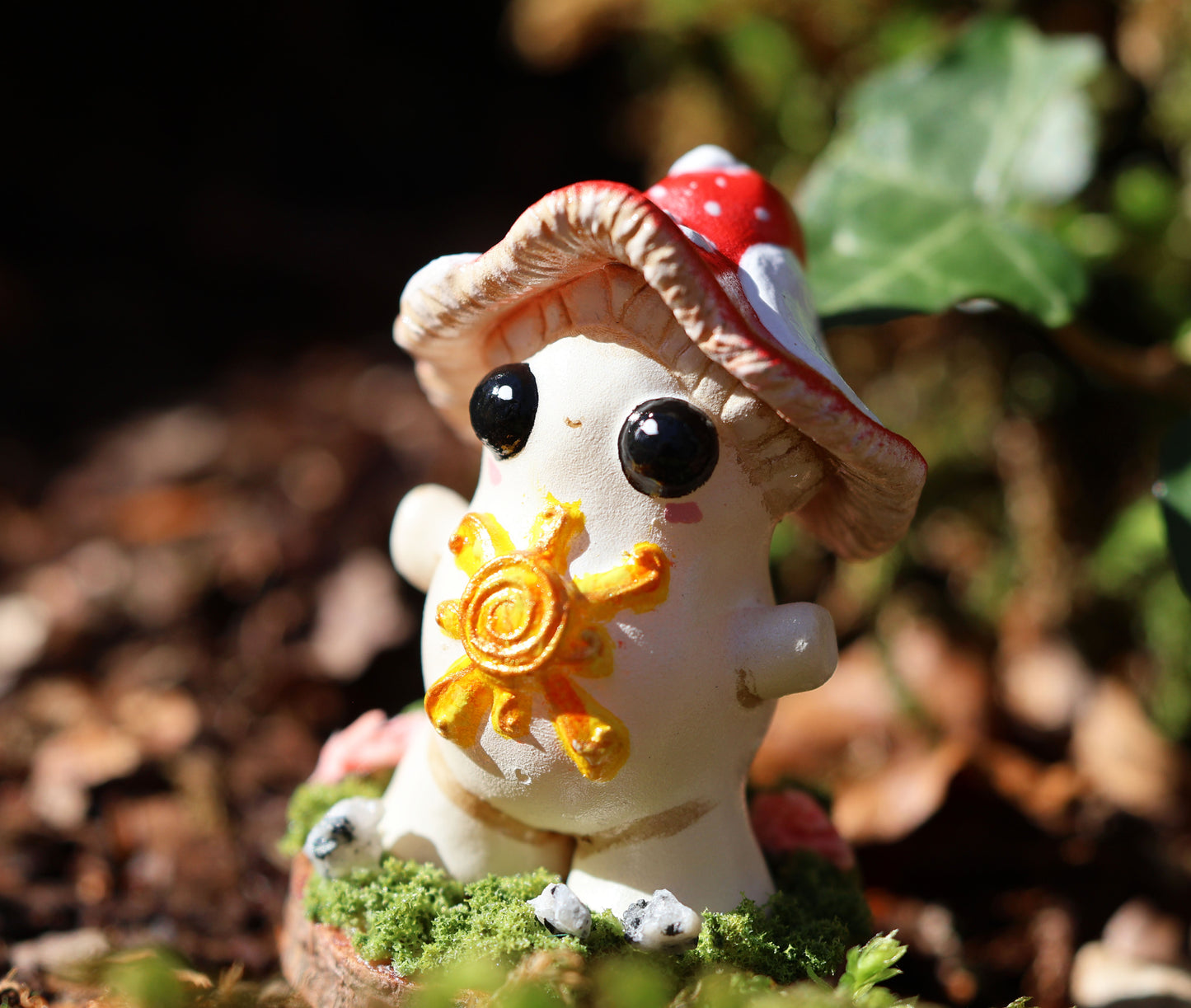 the photo shows alba, the light bringer, who is a fly agaric mushroom. she has a golden sunshine on her soft belly, big cute black eyes, a red and white spotted cap, and she stands with her arms outspread in a loving stance. she is attached to a circular wooden base which is decorated with decorative moss, and small stones. the sculpture is places amongst fallen brown leaves and ivy, which can be seen blurred in the background of the image