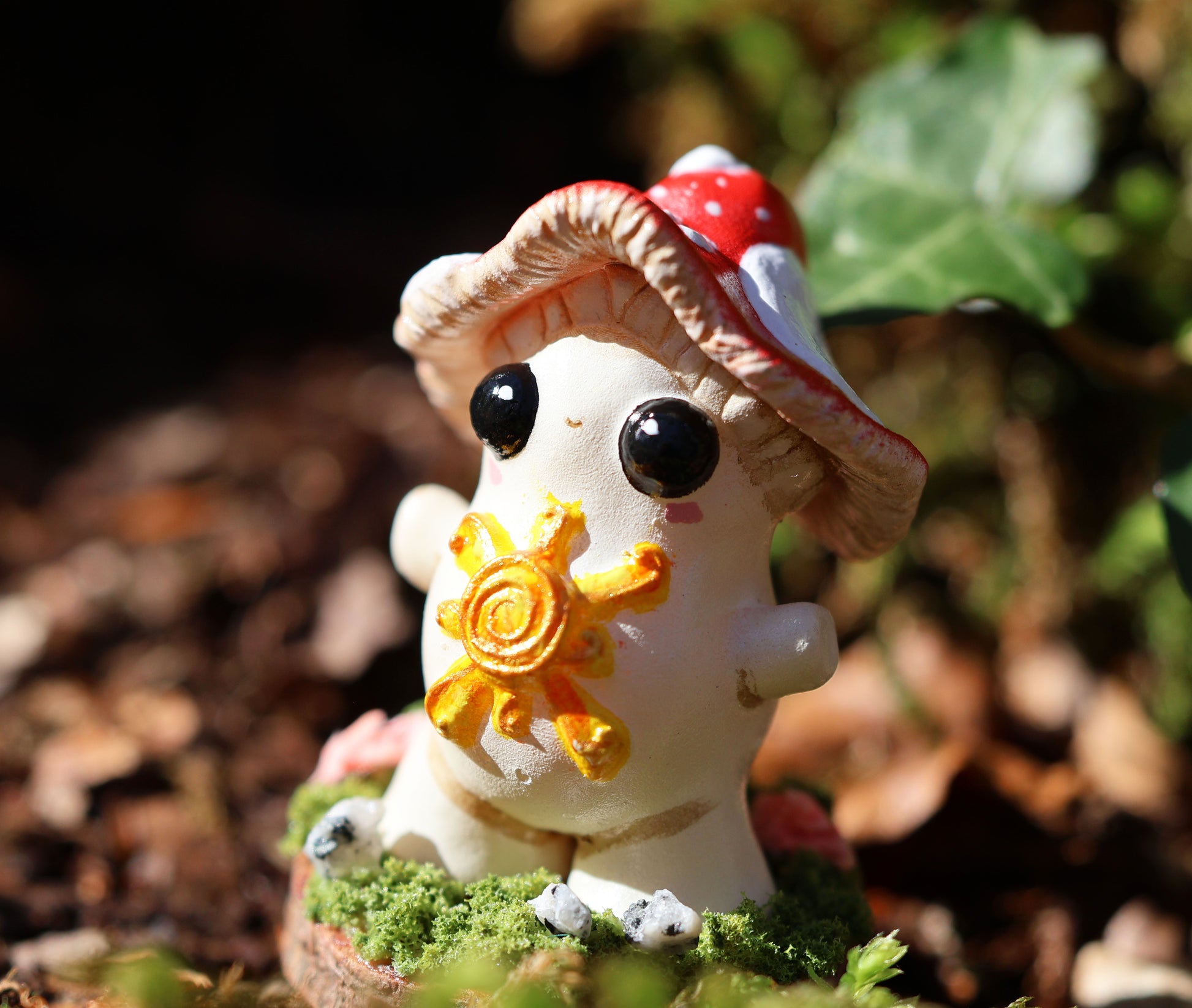 the photo shows alba, the light bringer, who is a fly agaric mushroom. she has a golden sunshine on her soft belly, big cute black eyes, a red and white spotted cap, and she stands with her arms outspread in a loving stance. she is attached to a circular wooden base which is decorated with decorative moss, and small stones. the sculpture is places amongst fallen brown leaves and ivy, which can be seen blurred in the background of the image