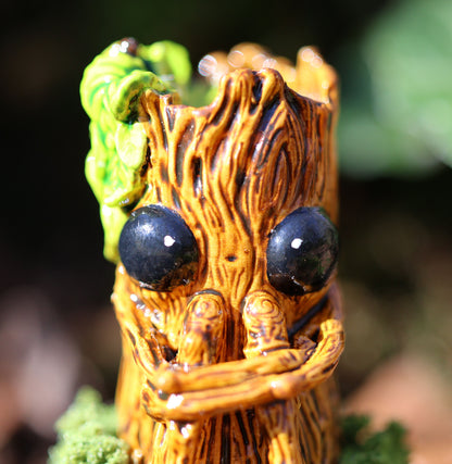 image shows a close up photo of splint the twigling, miniature sculpture. splint is seated on a muddy, mossy base, decorated with stones, he holds his legs close to his body, with a worried expression. he has big black eyes, and a bushell of green leaves coming from the left side of his head. 