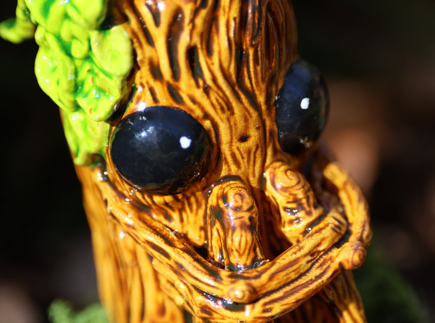 image shows a close up photo of splint the twigling, miniature sculpture .  he holds his legs close to his body, with a worried expression. he has big black eyes, and a bushell of green leaves coming from the left side of his head. 