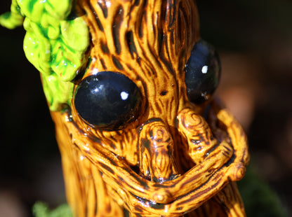 image shows a close up photo of splint the twigling, miniature sculpture .  he holds his legs close to his body, with a worried expression. he has big black eyes, and a bushell of green leaves coming from the left side of his head. 