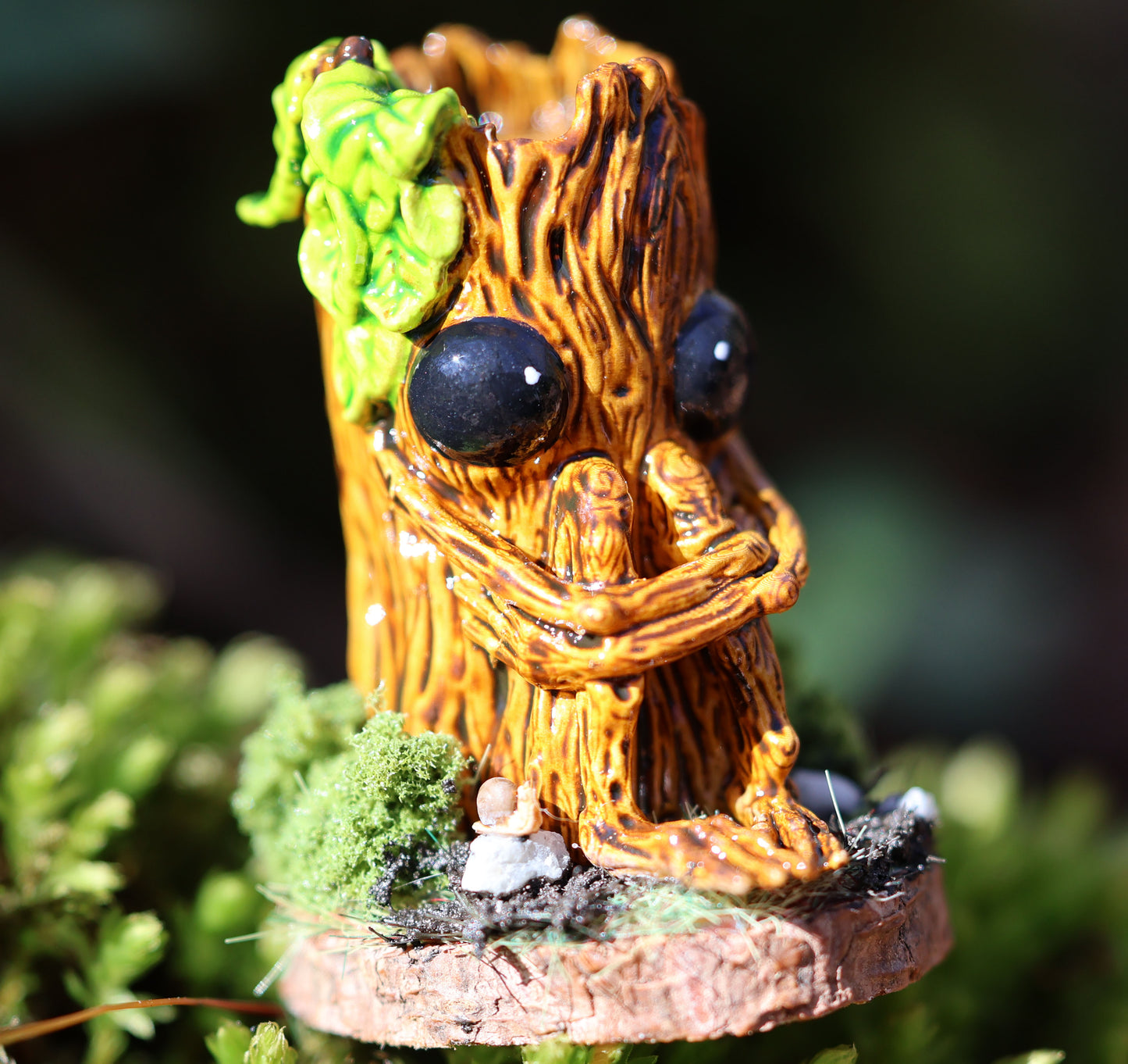 image shows a close up photo of splint the twigling, miniature sculpture being held between fingertips. splint is seated on a muddy, mossy base, decorated with stones, one of which features Splints little snail friend. he holds his legs close to his body, with a worried expression. he has big black eyes, and a bushell of green leaves coming from the left side of his head. 
