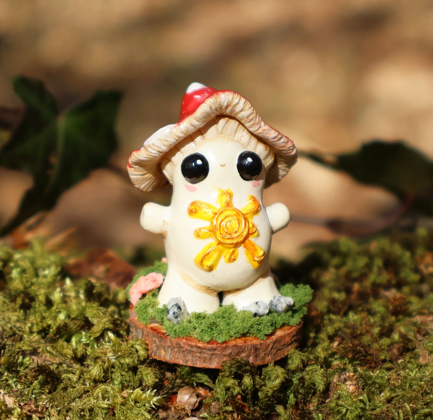 the photo shows alba, the light bringer, who is a fly agaric mushroom. she has a golden sunshine on her soft belly, big cute black eyes, a red and white spotted cap, and she stands with her arms outspread in a loving stance. she is attached to a circular wooden base which is decorated with decorative moss, and small stones. the sculpture is placed on a mossy log with ivy in the background