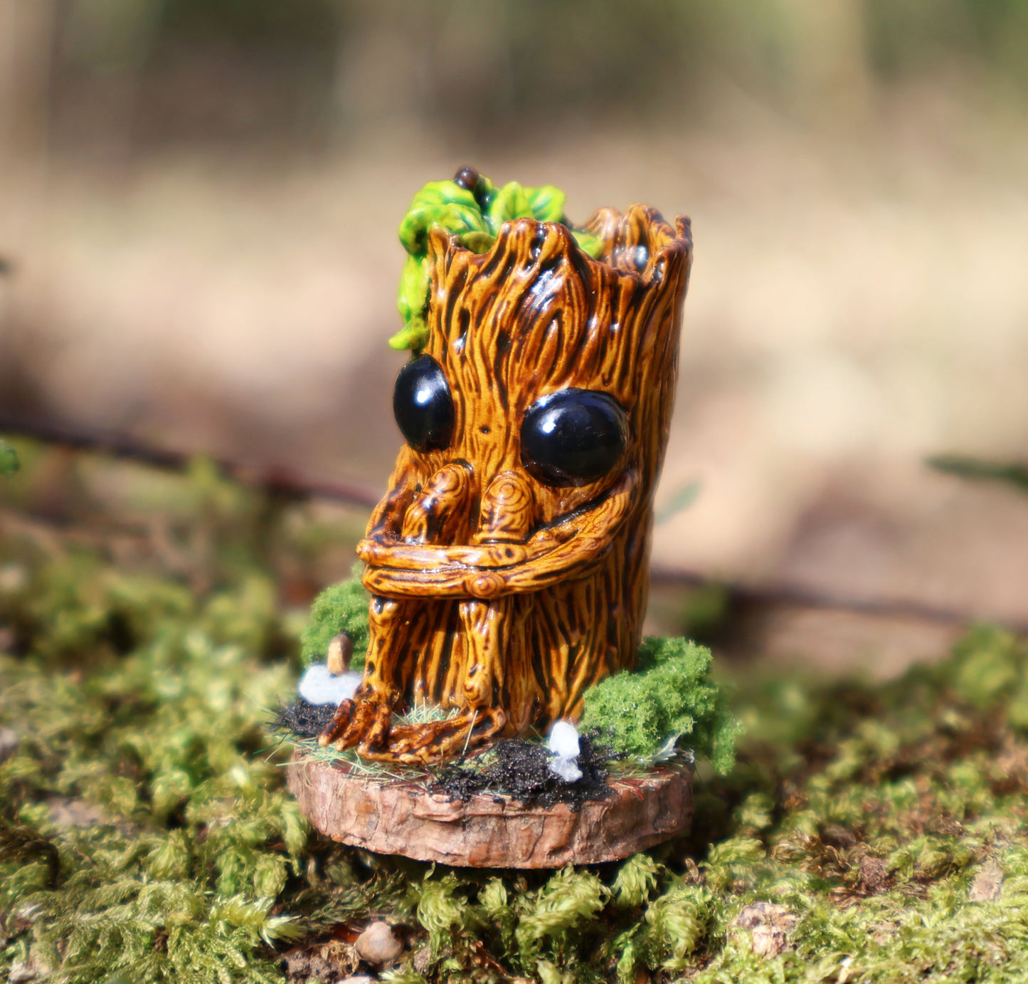 image shows a close up photo of splint the twigling, miniature sculpture being held between fingertips. splint is seated on a muddy, mossy base, decorated with stones, he holds his legs close to his body, with a worried expression. he has big black eyes, and a bushell of green leaves coming from the left side of his head. 