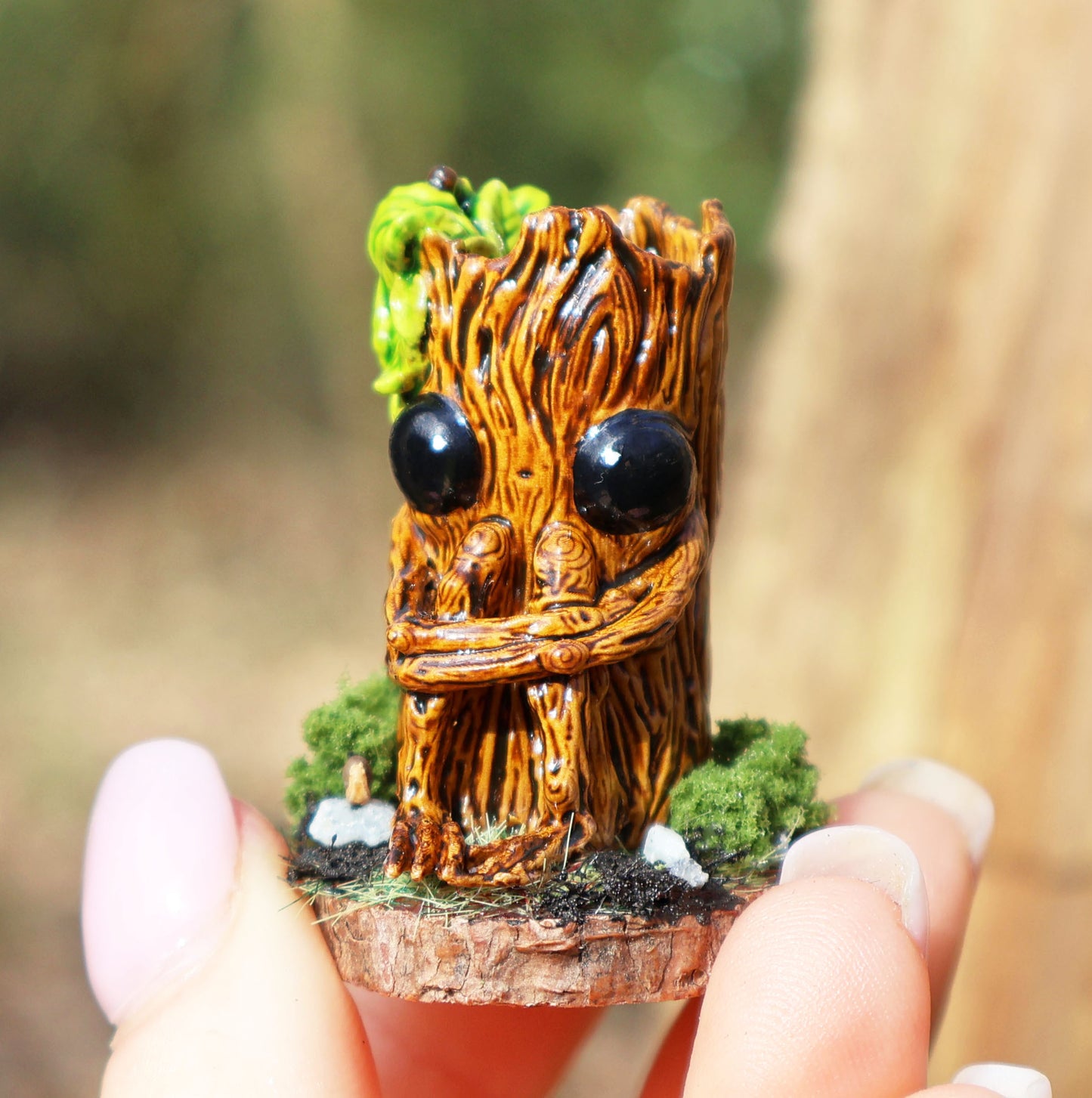 image shows a close up photo of splint the twigling, miniature sculpture being held between fingertips. splint is seated on a muddy, mossy base, decorated with stones, he holds his legs close to his body, with a worried expression. he has big black eyes, and a bushell of green leaves coming from the left side of his head. 