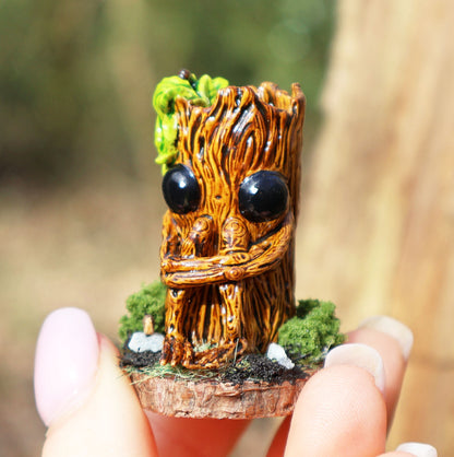 image shows a close up photo of splint the twigling, miniature sculpture being held between fingertips. splint is seated on a muddy, mossy base, decorated with stones, he holds his legs close to his body, with a worried expression. he has big black eyes, and a bushell of green leaves coming from the left side of his head. 
