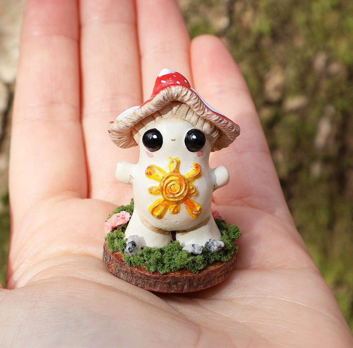 the photo shows alba, the light bringer, who is a fly agaric mushroom. she has a golden sunshine on her soft belly, big cute black eyes, a red and white spotted cap, and she stands with her arms outspread in a loving stance. she is attached to a circular wooden base which is decorated with decorative moss, and small stones. the sculpture is placed on the palm of an outstretched hand