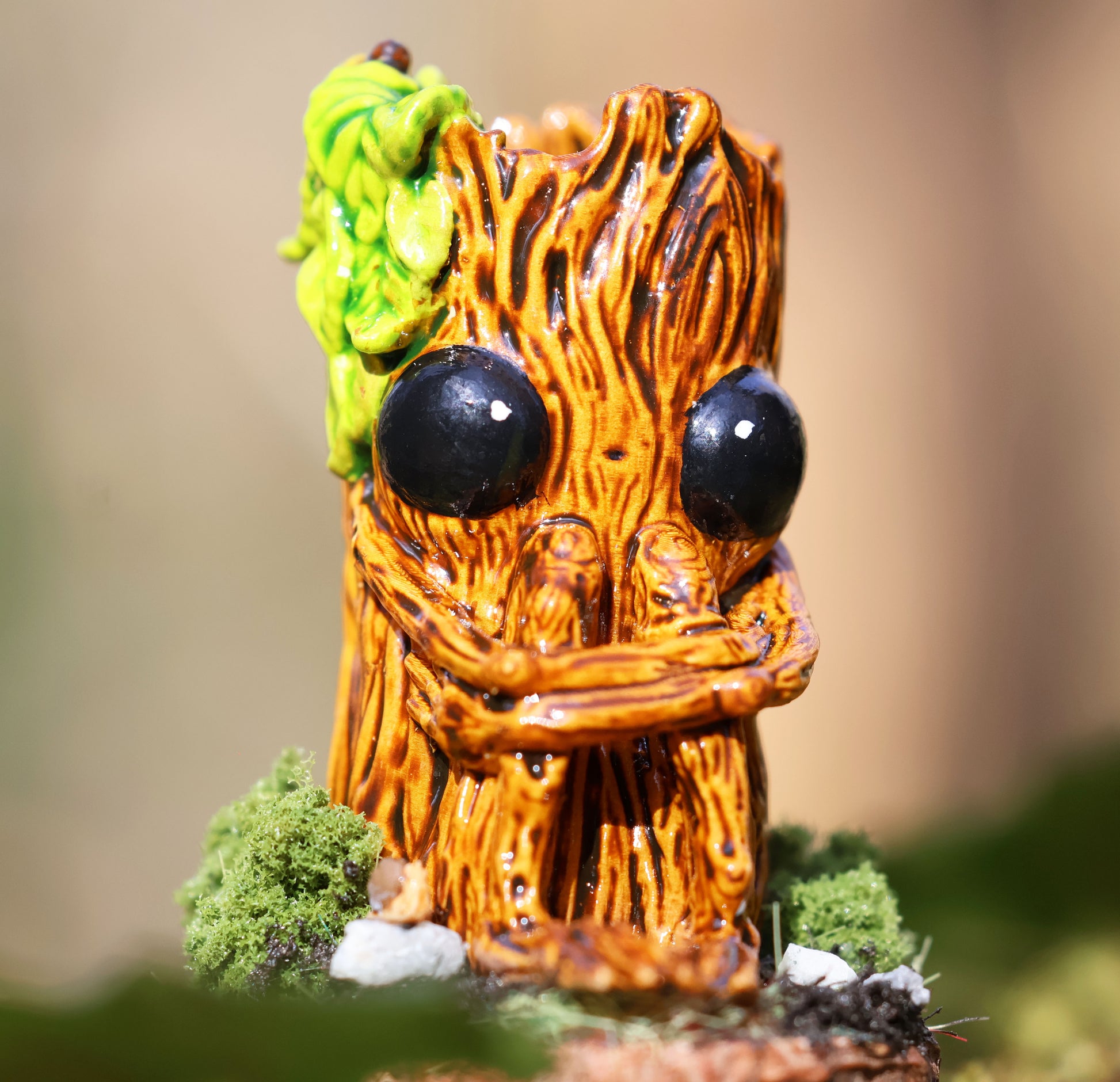image shows a close up photo of splint the twigling, miniature sculpture. splint is seated on a muddy, mossy base, decorated with stones, he holds his legs close to his body, with a worried expression. he has big black eyes, and a bushell of green leaves coming from the left side of his head. 