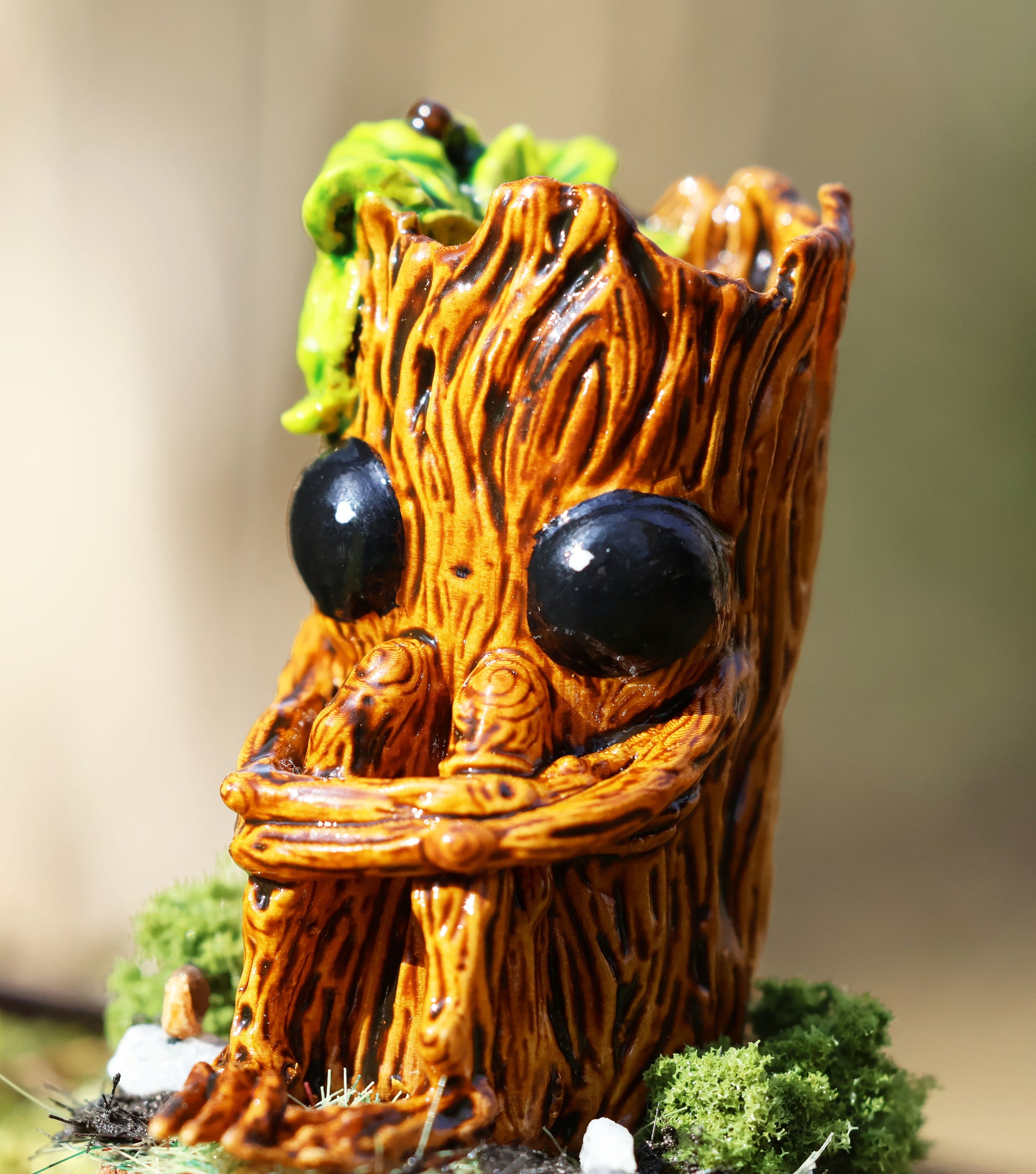 image shows a close up photo of splint the twigling, miniature sculpture. splint is seated on a muddy, mossy base, decorated with stones, he holds his legs close to his body, with a worried expression. he has big black eyes, and a bushell of green leaves coming from the left side of his head. 
