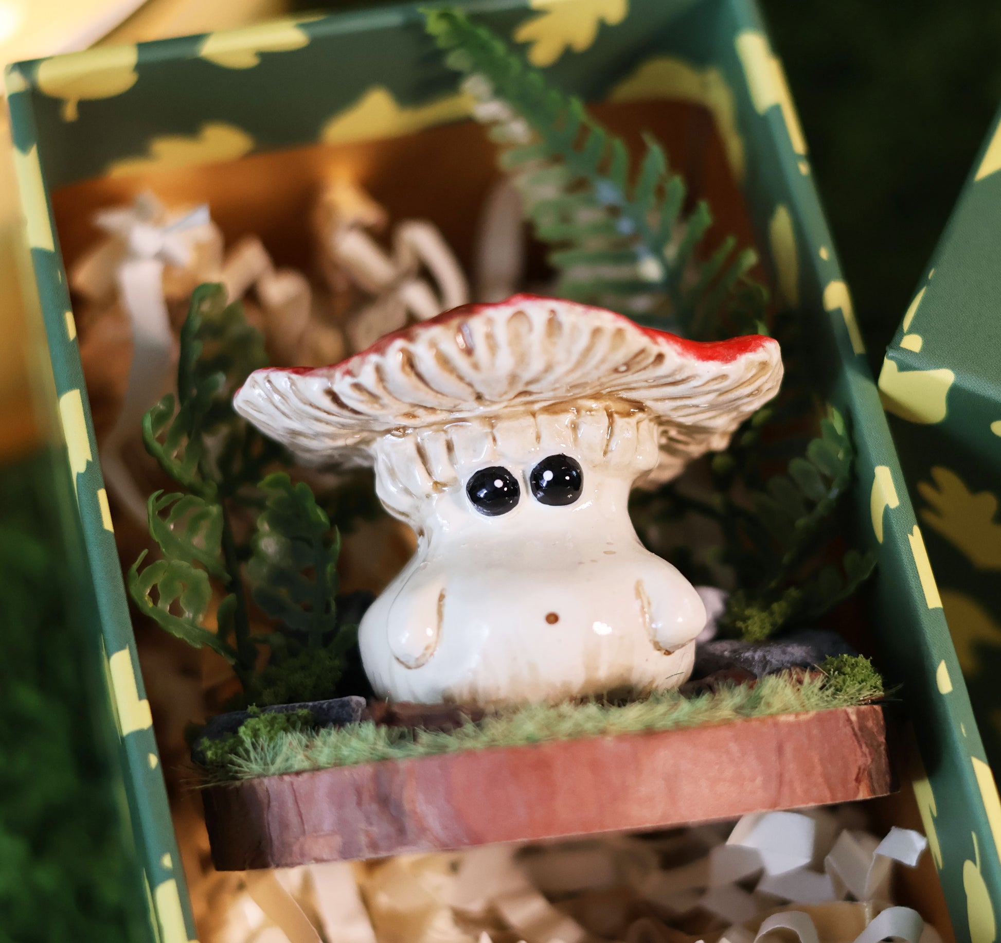 Adorable fly agaric mushroom figurine with a colourful red cap placed on a wooden base surrounded by a diorama of plants is placed inside a collectors display box filled with luxury packaging