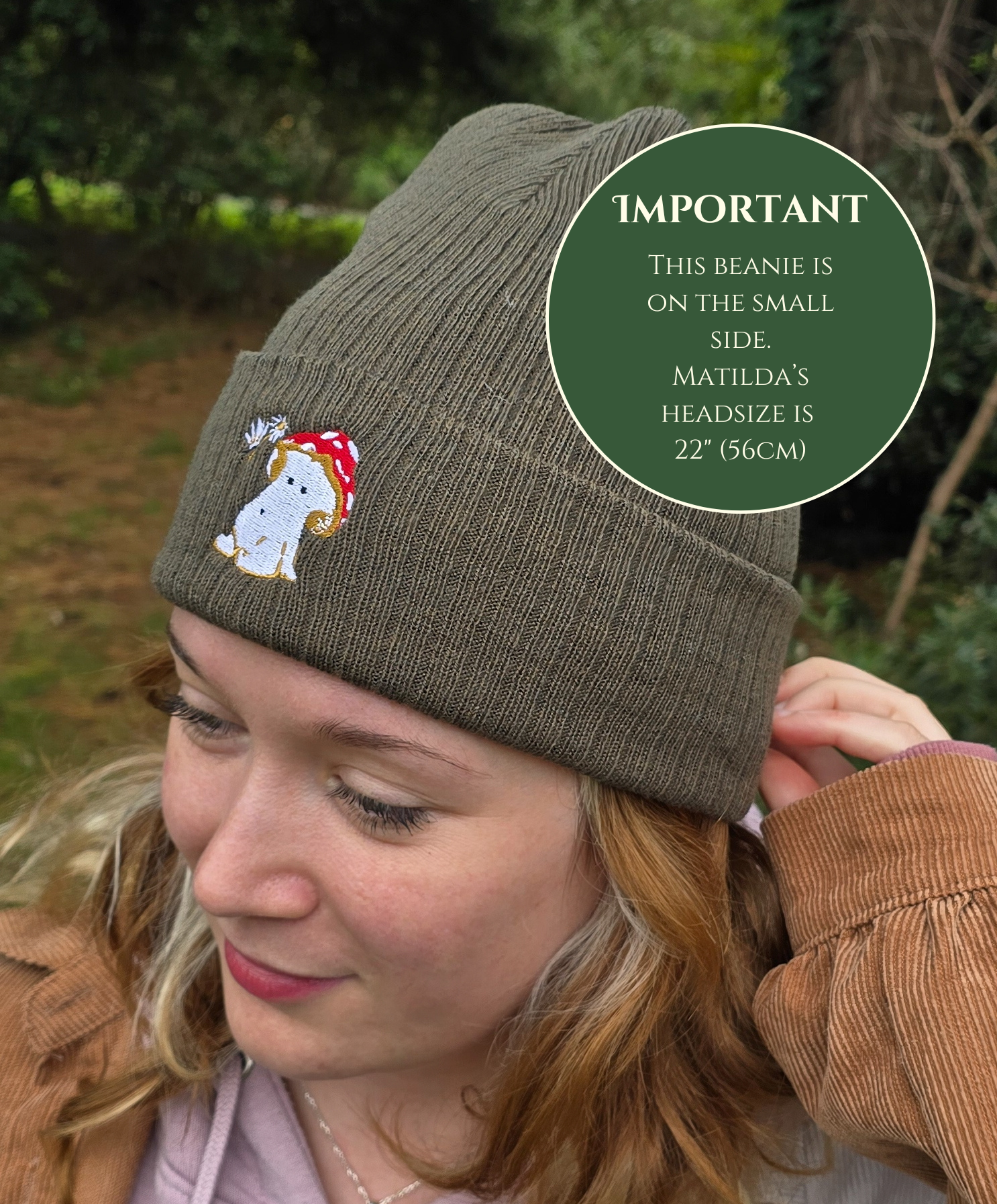 model wears a khaki green ribbed beanie hat with an embroidered image of a fly agaric toadstool mushroom seated, a fairy is beside the mushroom handing them a daisy

model is photographed in front of trees in a forest

text explains beanie hat comes up small, models head size is 22" / 56cm