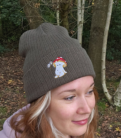 model wears a khaki green ribbed beanie hat with an embroidered image of a fly agaric toadstool mushroom seated, a fairy is beside the mushroom handing them a daisy

model is photographed in front of trees in a forest