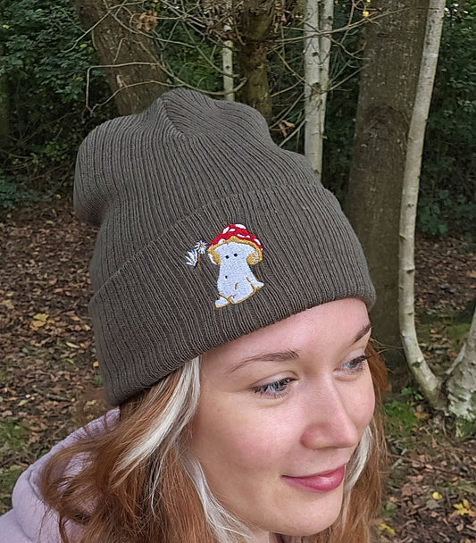 model wears a khaki green ribbed beanie hat with an embroidered image of a fly agaric toadstool mushroom seated, a fairy is beside the mushroom handing them a daisy

model is photographed in front of trees in a forest