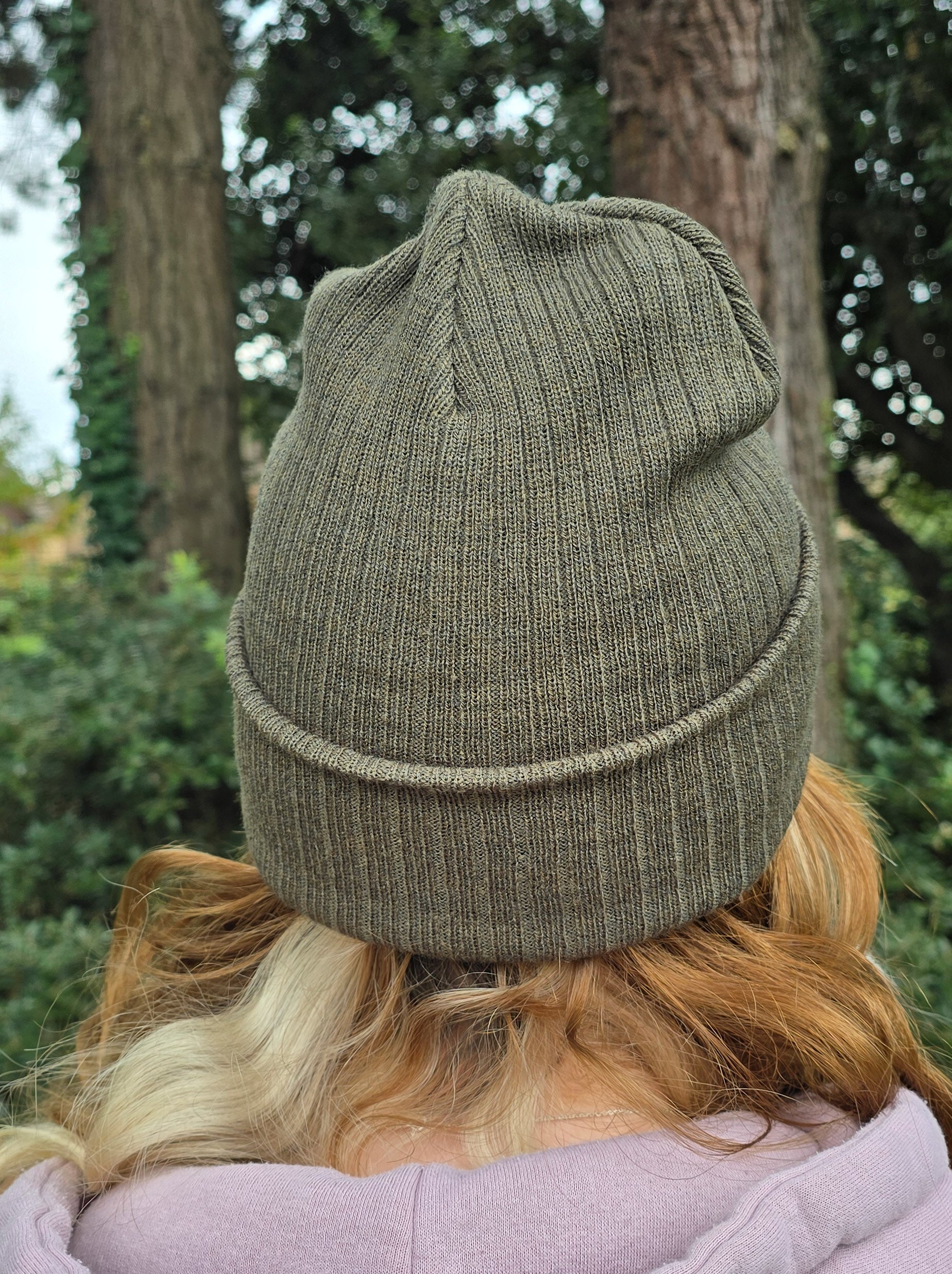 model wears a khaki green ribbed beanie hat, showing the back of the hat 

photographed in front of ivy covered trees in a forest