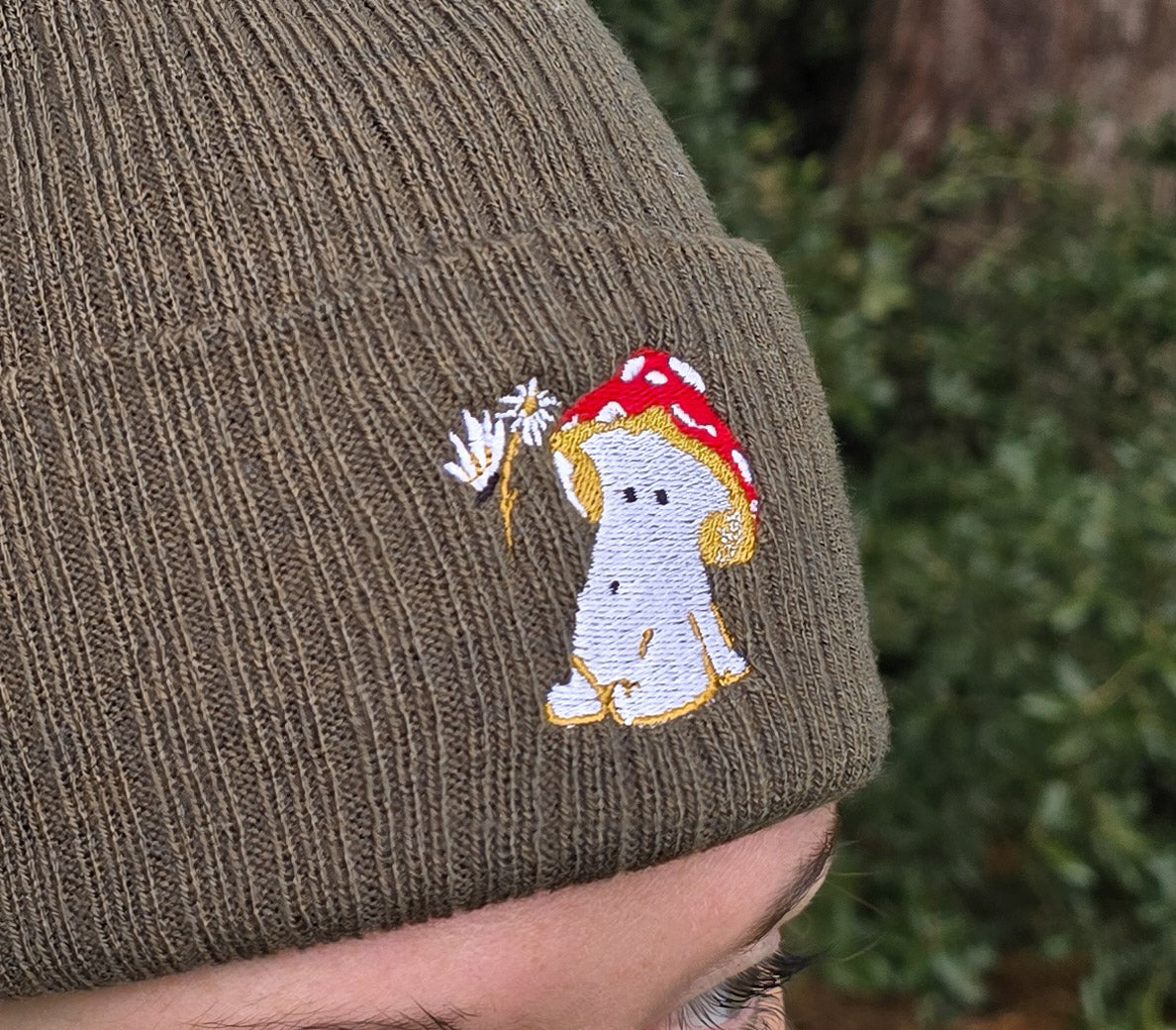 model wears a khaki green ribbed beanie hat with an embroidered image of a fly agaric toadstool mushroom seated, a fairy is beside the mushroom handing them a daisy

model is photographed in front of trees in a forest