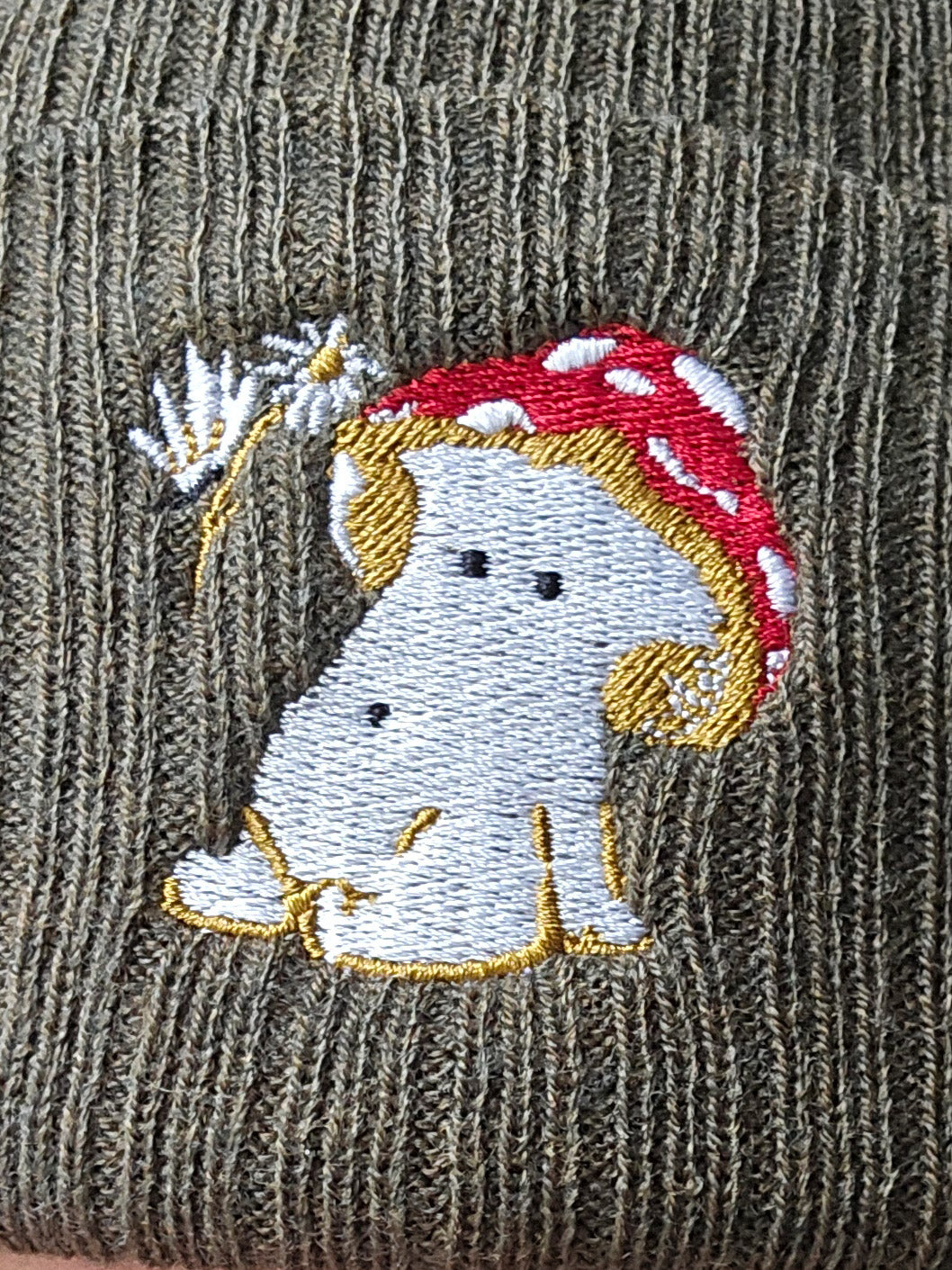 a close up photo of the embroidered image of a fly agaric toadstool mushroom seated, a fairy is beside the mushroom handing them a daisy, placed onto a khaki green ribbed beanie hat