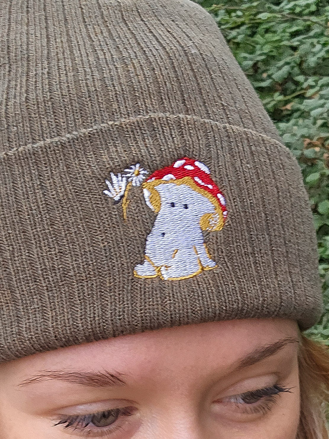 model wears a khaki green ribbed beanie hat with an embroidered image of a fly agaric toadstool mushroom seated, a fairy is beside the mushroom handing them a daisy

model is photographed in front of trees in a forest