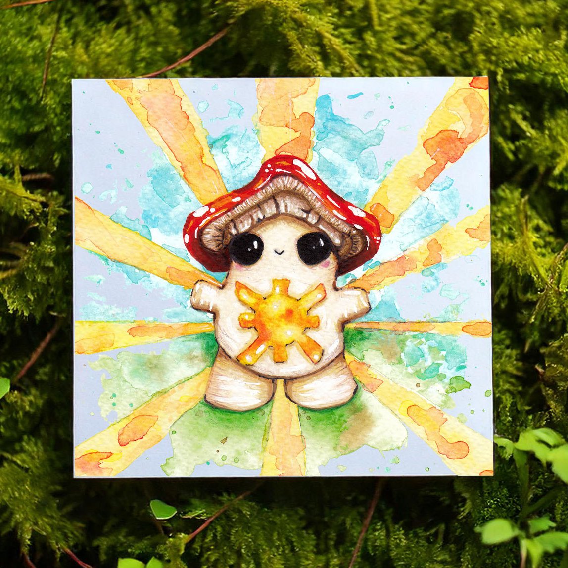 the photo shows an art print laying on top of a mossy ground. The art print features a smiley toadstool mushroom emitting a sunny glow from her belly. around her are blue and green watercolour markings