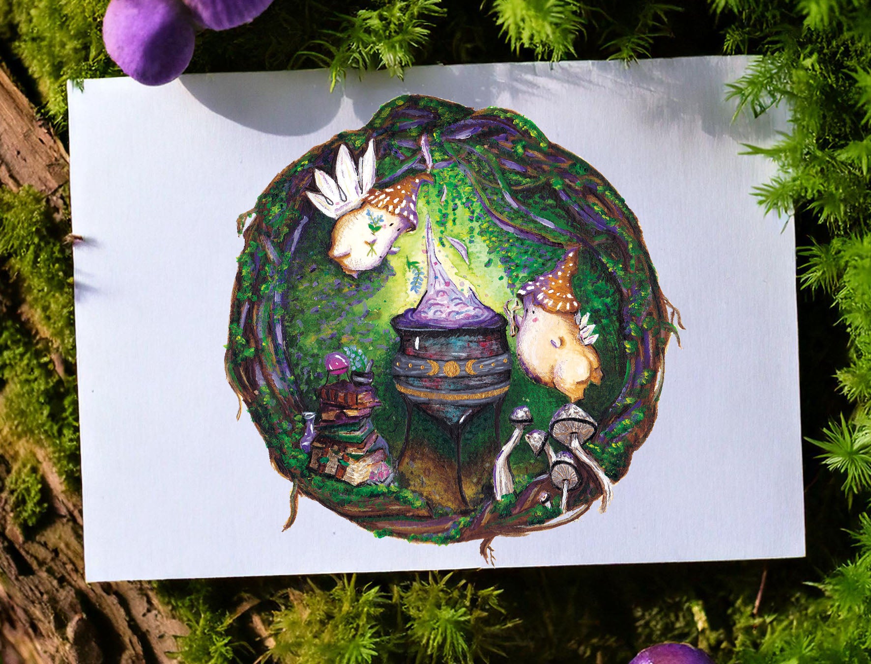 An art print with magical witch mushroom folk around a cauldron brewing a purple glowing potion is resting on a mossy rock. The art print rests on a moss covered tree stump with mushrooms growing from the moss