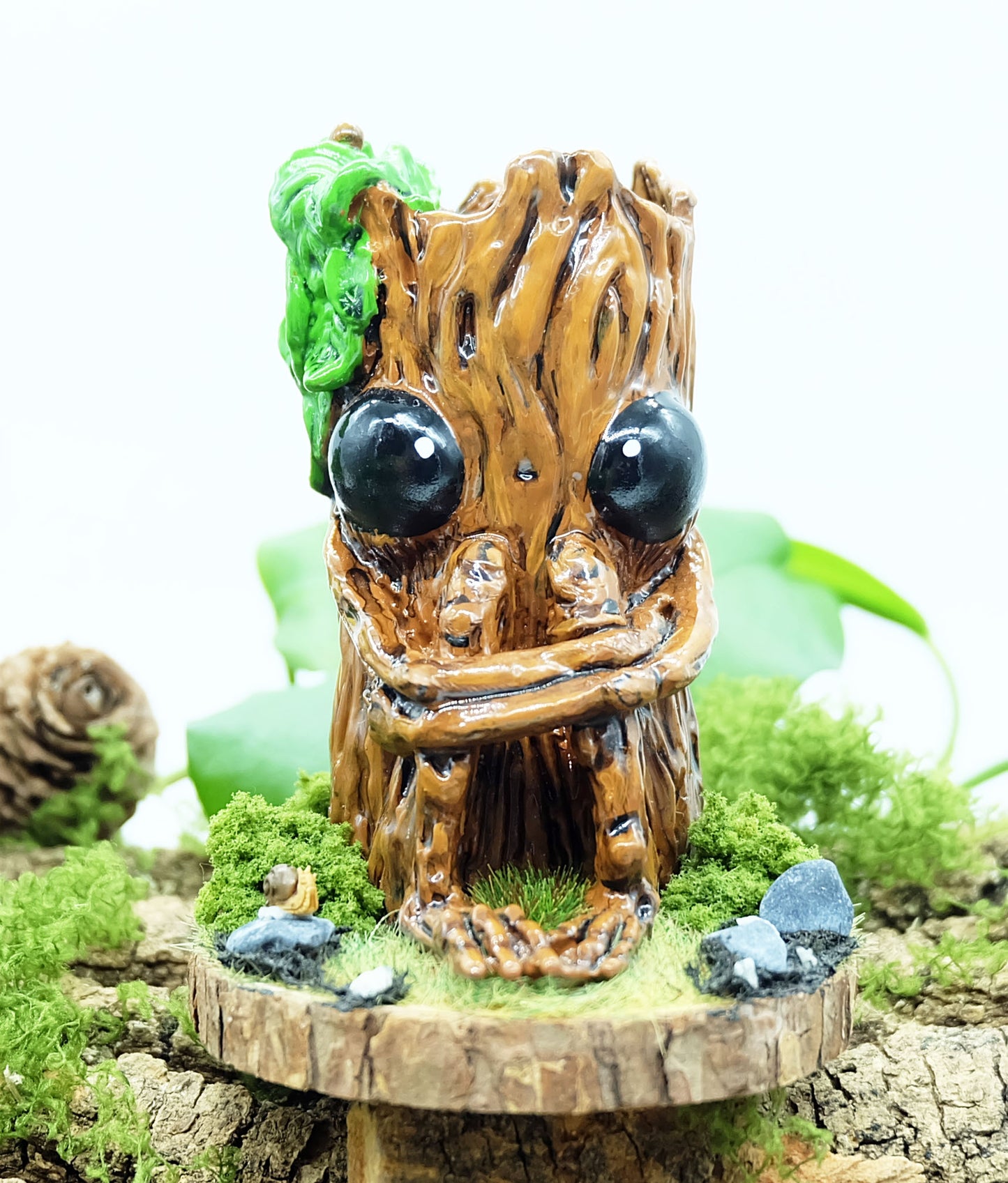 hand made sculpture of Splint the Twigling, a tree stump with big black eyes and an anxious expression hugs their knees as they are seated upon a wooden disc, surrounded by moss, grass, rocks and a tiny snail friend. Splint has green leaves coming from the top of their head, and is photographed against a white background with leaves and plants behind the sculpture