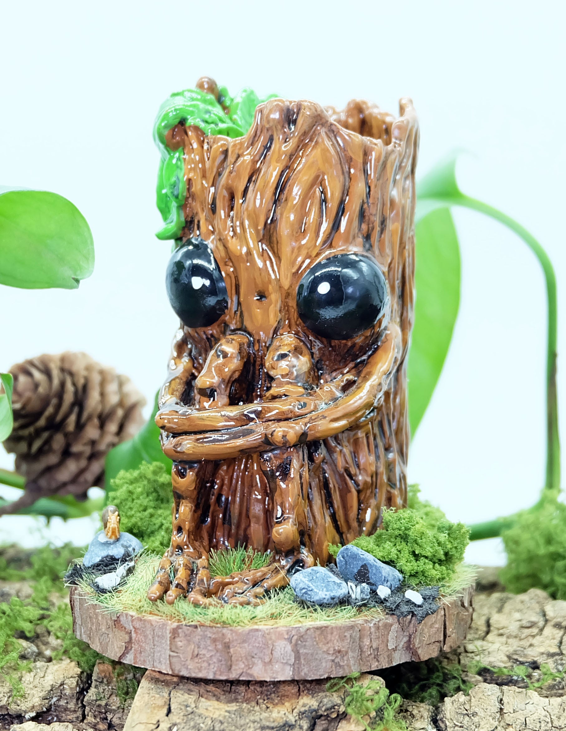 hand made sculpture of Splint the Twigling, a tree stump with big black eyes and an anxious expression hugs their knees as they are seated upon a wooden disc, surrounded by moss, grass, rocks and a tiny snail friend. Splint has green leaves coming from the top of their head, and is photographed against a white background with leaves and plants behind the sculpture